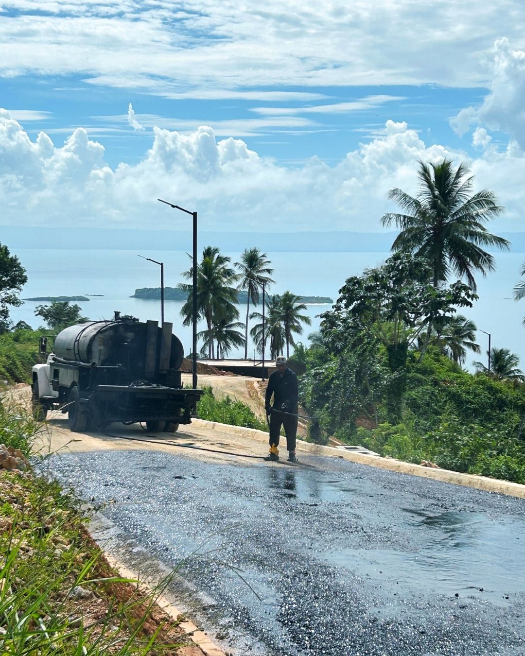 Ouvrier appliquant une couche de liant sur la surface de la route interne à Nomad City 1A, avec un camion-citerne et la baie de Samaná en arrière-plan.