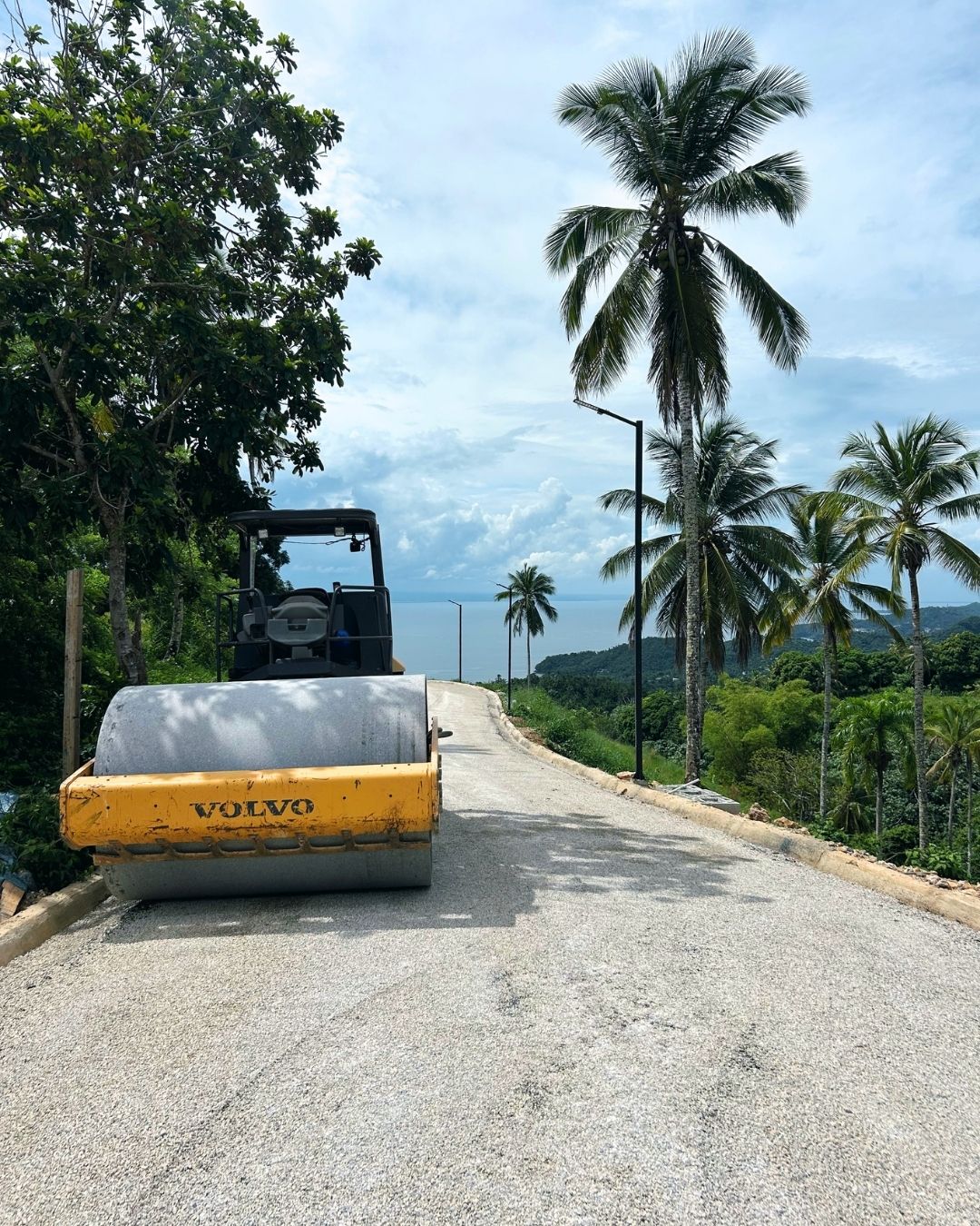 Rouleau compresseur compactant la surface d'agrégats fraîchement posée sur une route interne à Nomad City 1A, entourée de végétation tropicale.