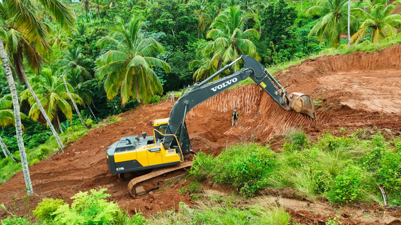 An excavator cutting a terrace into red earth on a palm-covered hillside