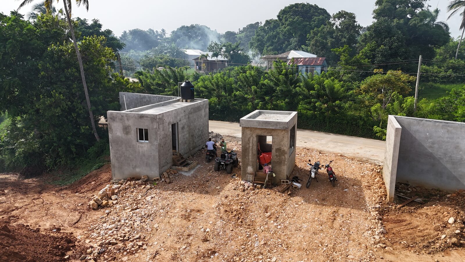 Two concrete structures with block walls standing beside a newly cut earth road, workers and motorcycles in the foreground