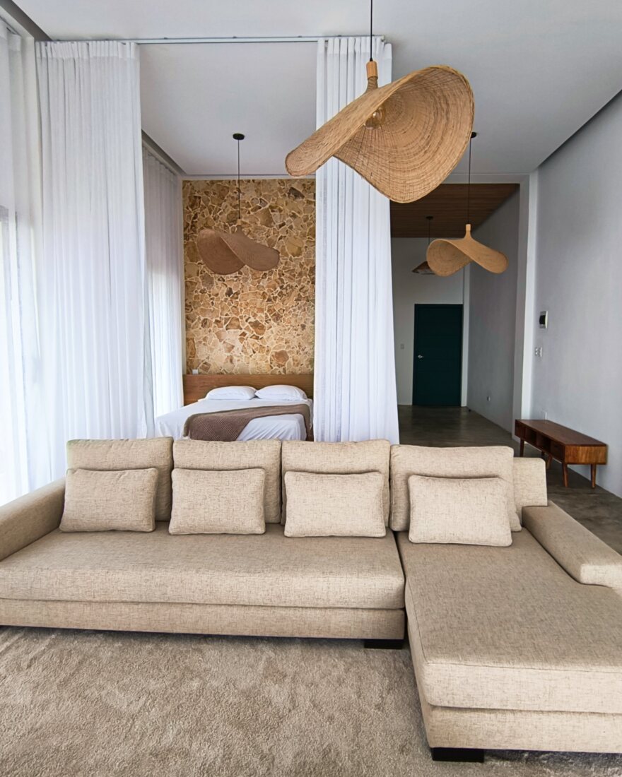 Showroom living room with a low linen sofa, rattan pendant lights and a bedroom visible beyond