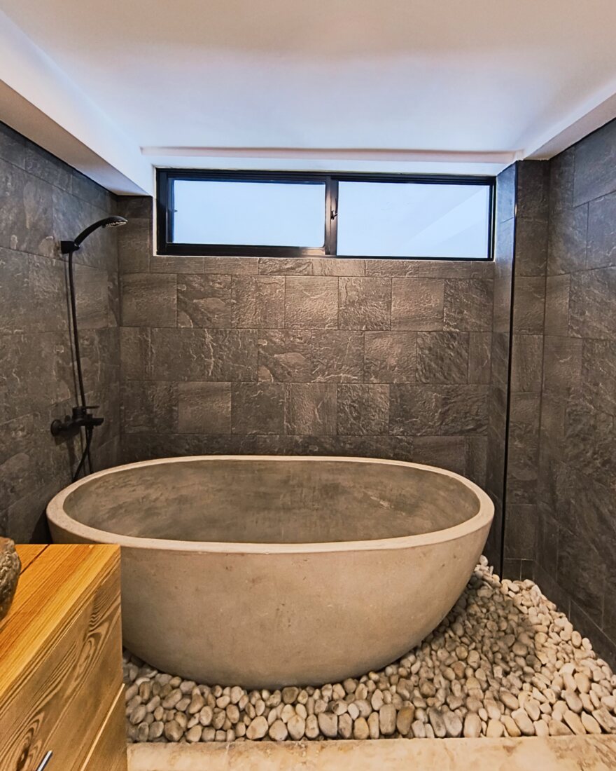 A rounded concrete soaking tub set on river stones beneath a window