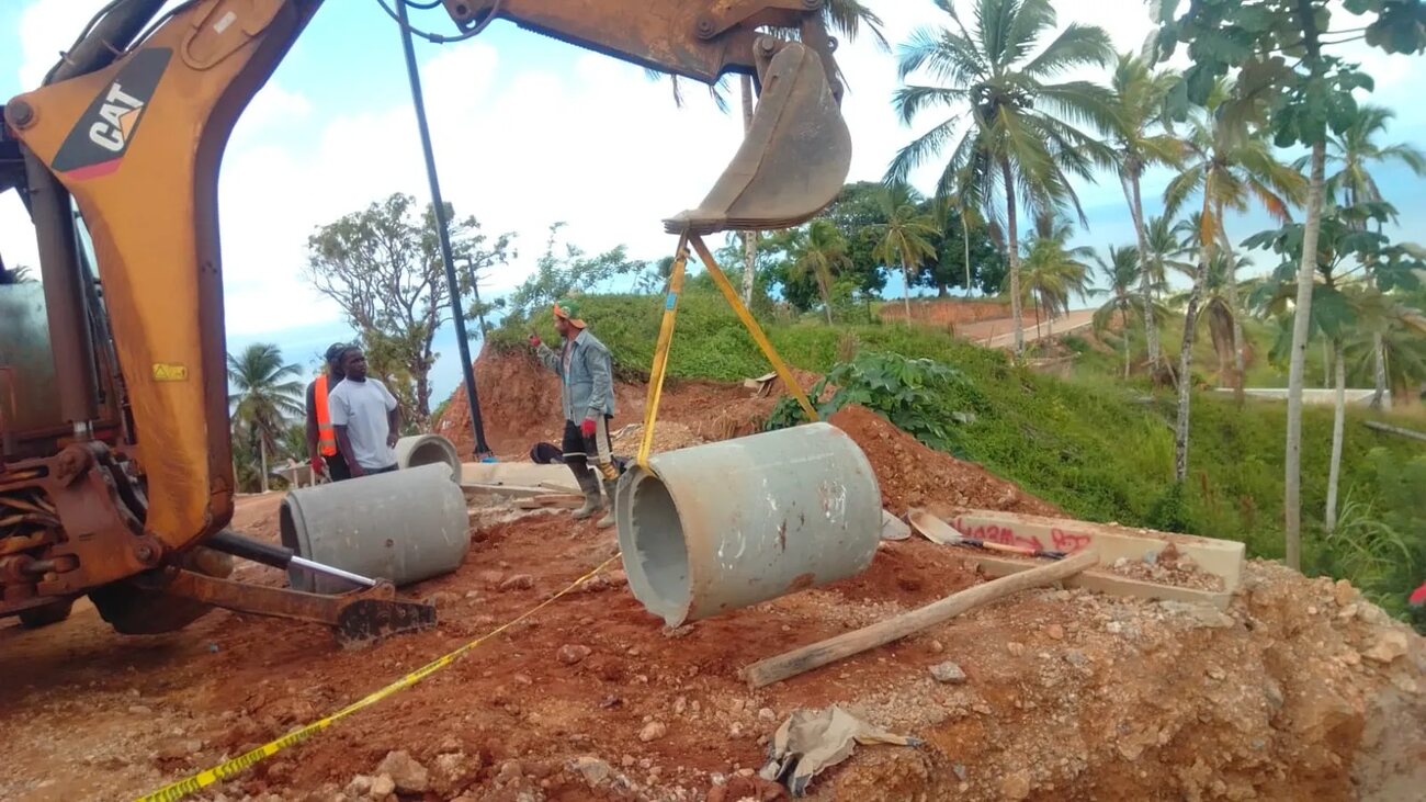An excavator lowering a concrete pipe on slings while three workers guide it into position on red earth