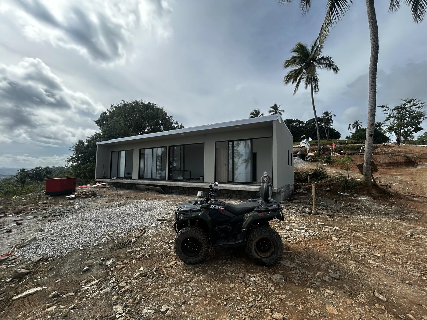 A completed single-storey Flat House with full glazing, an ATV parked outside, palms and sky behind