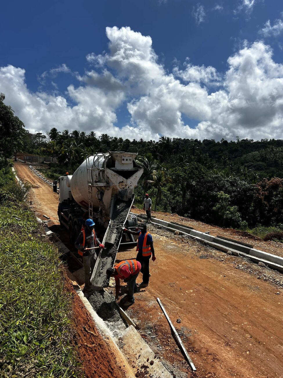 A concrete mixer truck discharging on a newly formed road cut through green vegetation
