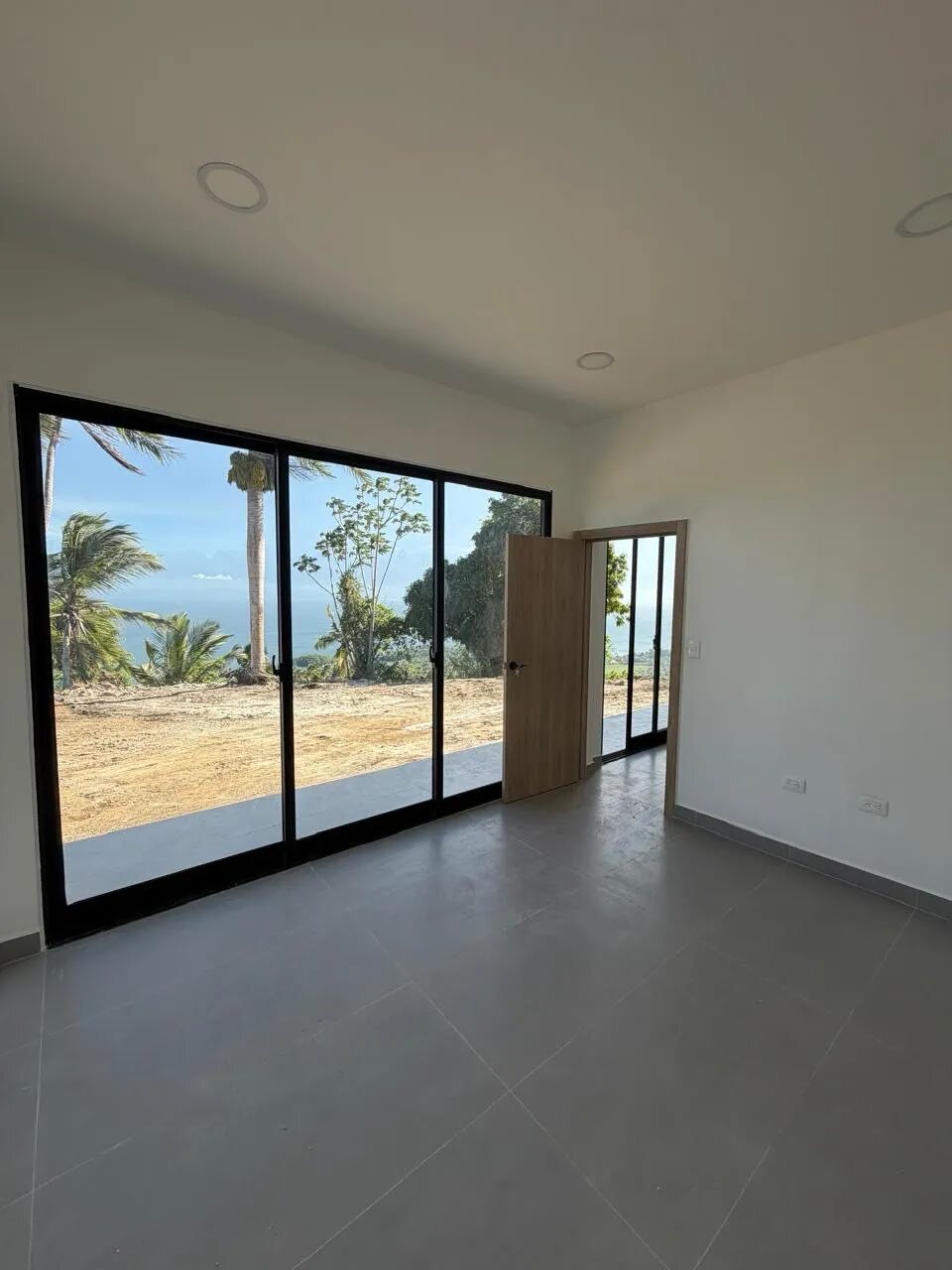 A bedroom with a wooden door open and full-height glazing framing palms and the sea