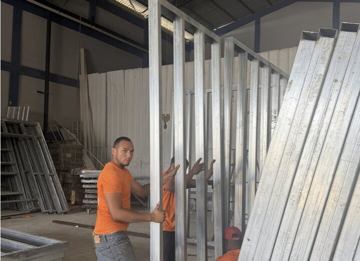 Two workers in orange shirts guiding a tall galvanised steel wall panel into position inside the fabrication plant