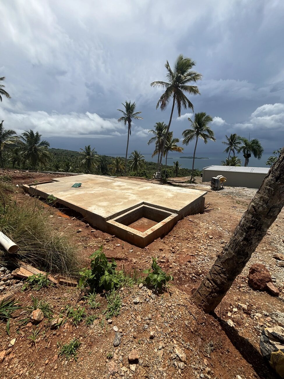 A poured house slab among coconut palms under a heavy sky, the sea and a headland in the distance