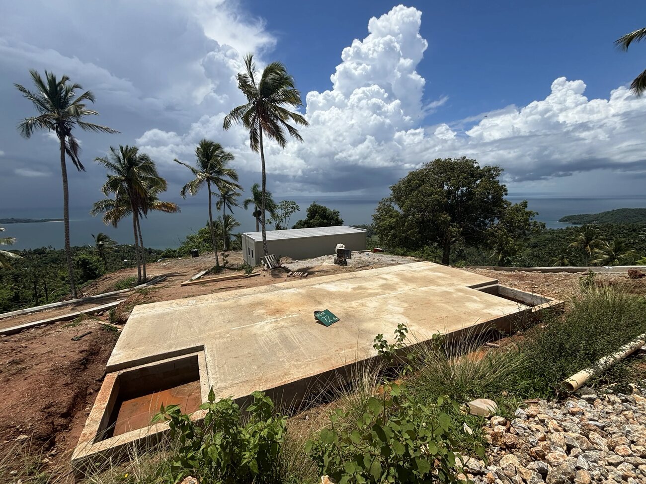 A newly poured concrete house slab on a hillside terrace with palms and Samana Bay beyond, a completed house in the background