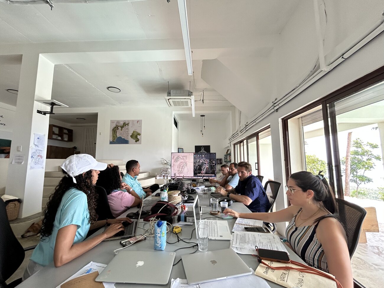 A long shared desk of people working on laptops in a bright site office, the Nomad City masterplan displayed on a large screen