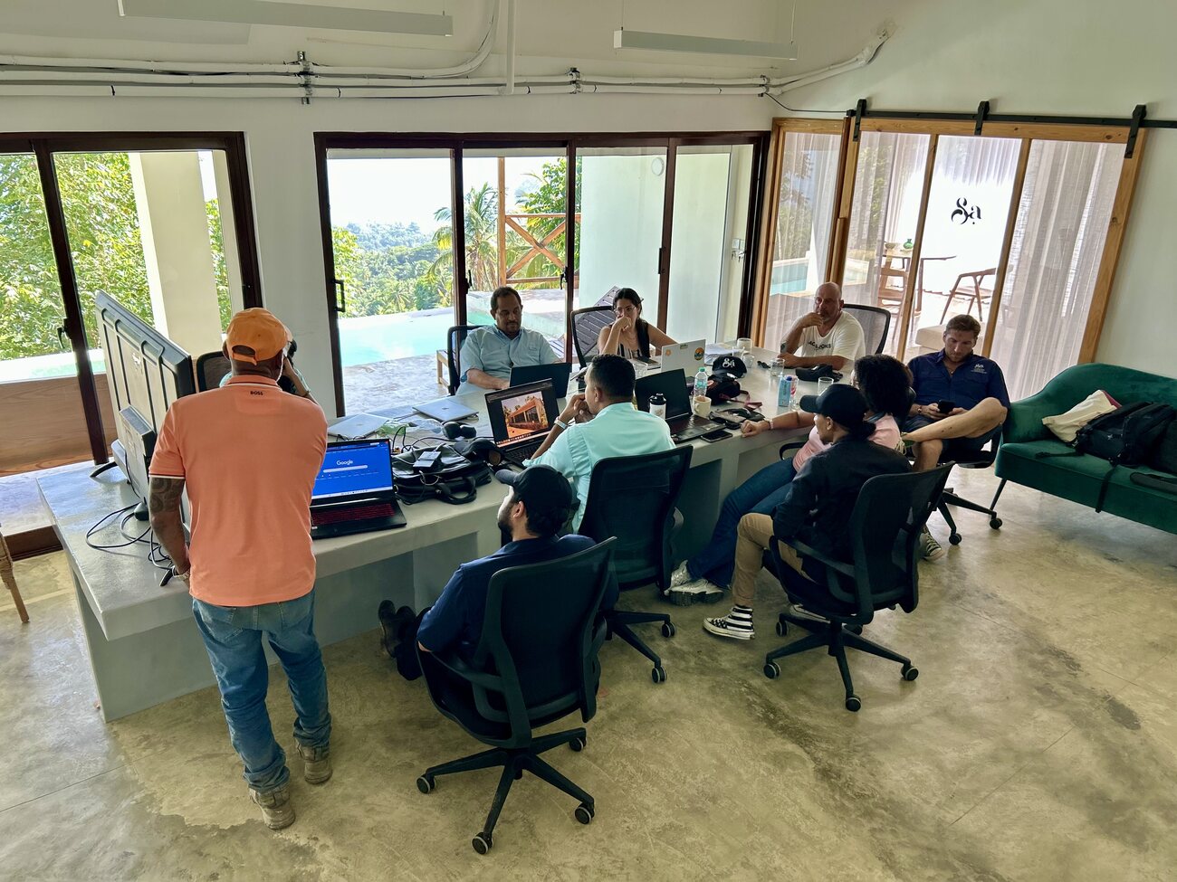 A dozen people seated around a large table in an open site office reviewing drawings and laptops, pool and forest beyond the glass